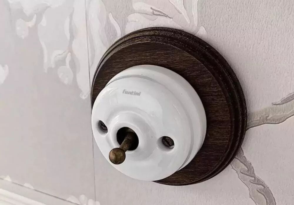 Installation d'un interrupteur en porcelaine blanche et levier et vis en laiton veiilli. L'interrupteur est pos� sur un socle en h�tre vieilli sur un mur � la tapisserie grise.
