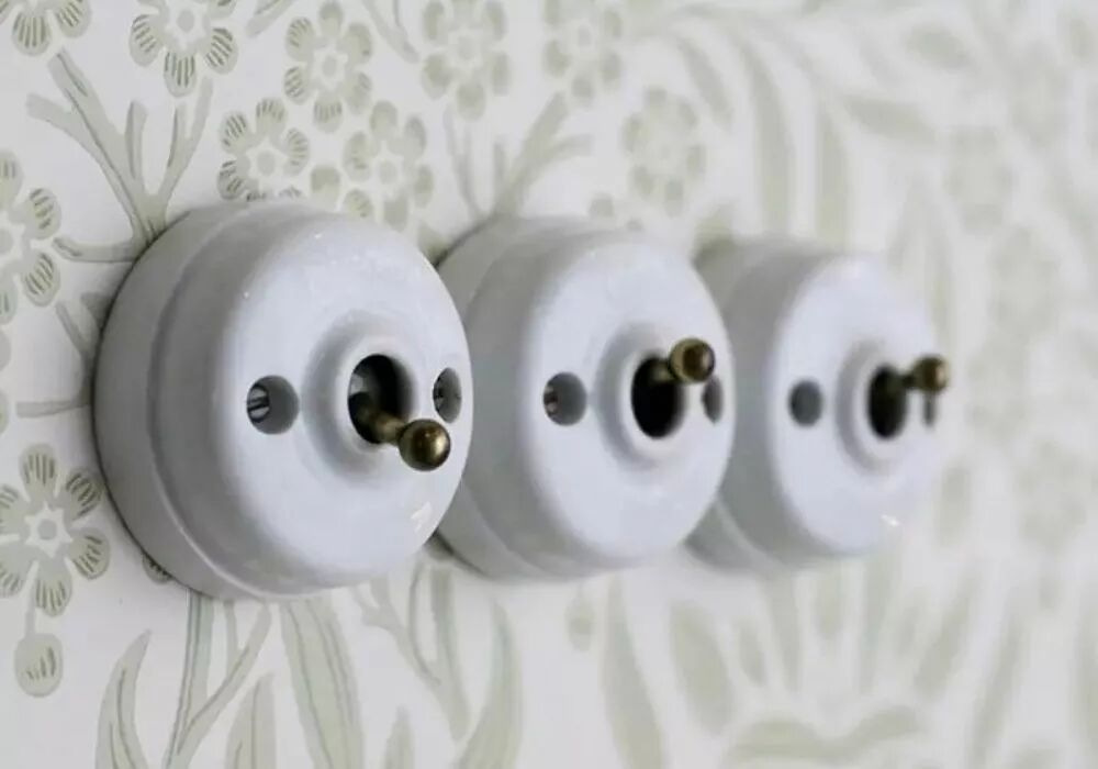Pr�sentation en saillie de trois interrupteurs Dimbler align�s. Leur base est en porcelaine blanche et leurs leviers sont en laiton vieilli. La tapisserie du mur pr�sente des motifs � fleurs.  