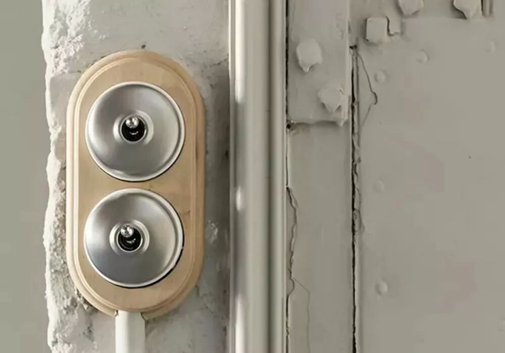Installation d'un socle double en h�tre verni avec deux interrupteurs en porcelaine blanche et d�me et levier chrom�s. Le mur blanc est d�cr�pi. 