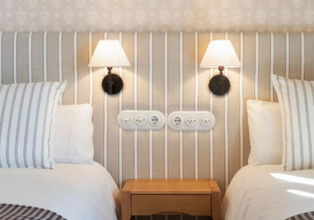 L'image sym�trique met en sc�ne deux plaques triples en porcelaine au-dessus d'une table de chevet. Ces plaques, dispos�es horizontalement, accueillent chacune une prise et deux interrupteurs en porcelaine blanche de la gamme Garby Colonial. Deux lits sont de chaque c�t� de la table de chevet dans cette chambre.