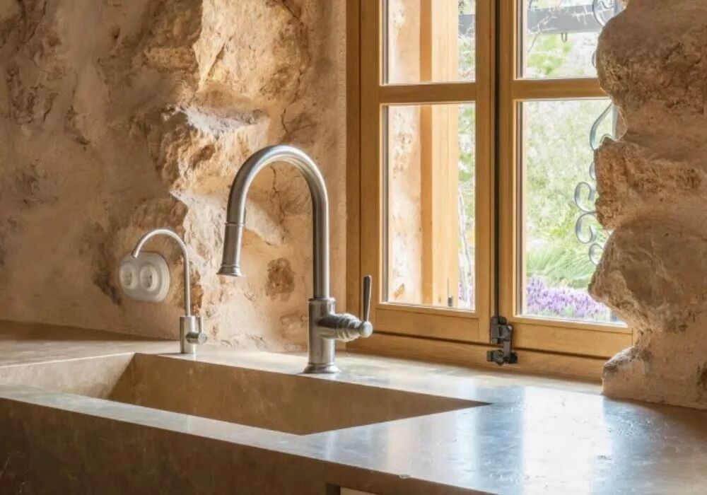 Dans une cuisine en pierre, alors qu'un �vier et son robinet profitent de la lumi�re glissant � travers la fen�tre qui leur fait face, une plaque double et ses interrupteurs en porcelaine blanche de la gamme Garby Colonial d�tachent leur silhouette du mur.