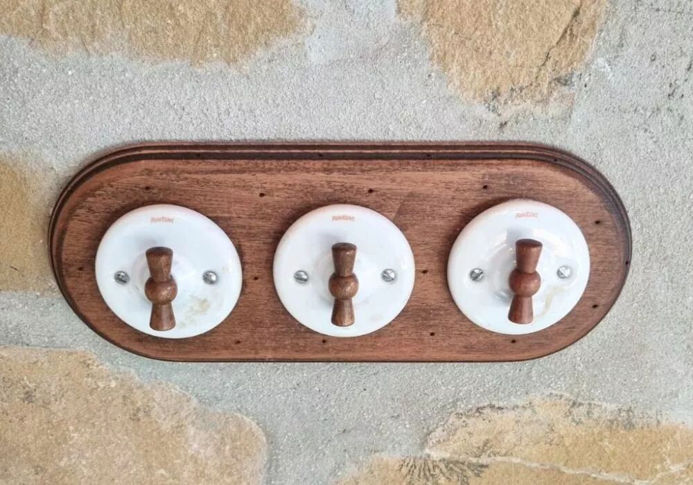 Sur un mur en pierre, une plaque triple en h�tre vieilli de la collection Garby Colonial accueille trois interrupteurs en porcelaine blanche � manette rotative en h�tre vieilli.