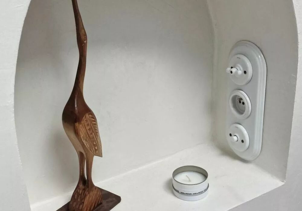 Dans une niche peinte en blanc, sur une surface lisse, se dessine une plaque de finition triple Garby Colonial. Install�e � l'horizontal, elle accueille un interrupteur � manette rotative en porcelaine, une prise de courant et un bouton poussoir de la m�me mati�re. Face � cette plaque, sur la niche, une bougie et une statue en bois d'oiseau sont pos�es.