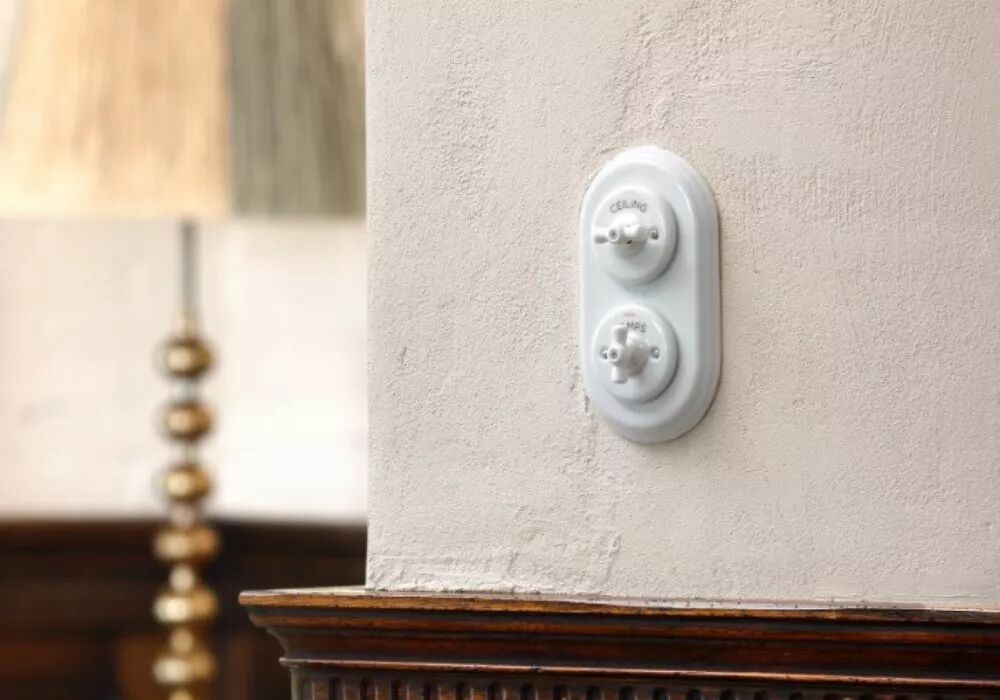 Sur un mur en cr�pit dont la base en bois se laisse entrevoir en bas de l'image, une plaque double en porcelaine blanche est install�e � la verticale. Cette plaque accueille deux interrupteurs � manette rotative en porcelaine blanche de la gamme Garby Colonial. 