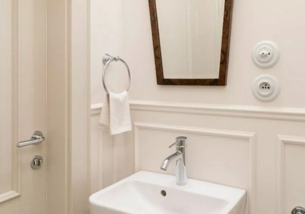 Dans une salle de bain avec moulures, au-dessus d'une vasque et � droite d'un miroir au cadre en bois, se trouvent un interrupteur et une prise de courant en porcelaine blanche. Chaque �l�ment est situ� dans une plaque en porcelaine blanche.