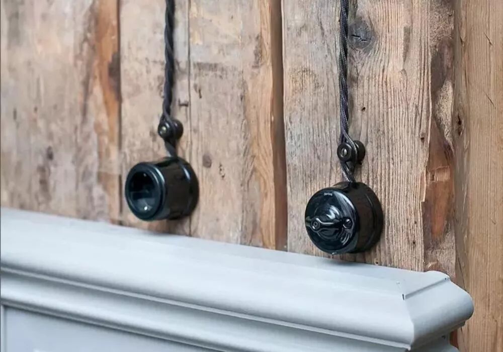 Dispos�s sur une surface en planches de bois, deux �l�ments Garby en porcelaine noire sont dispos�s. A gauche, une prise de courant est reli�e vers le haut par le c�ble. A droite, c'est un interrupteur rotatif qui dispose de la m�me configuration. 