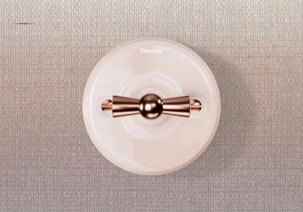 Sur un fond textile aux couleurs ros�es, un interrupteur Garby en porcelaine rose � manette rotative cuivr�e se distingue par sa silhouette r�tro